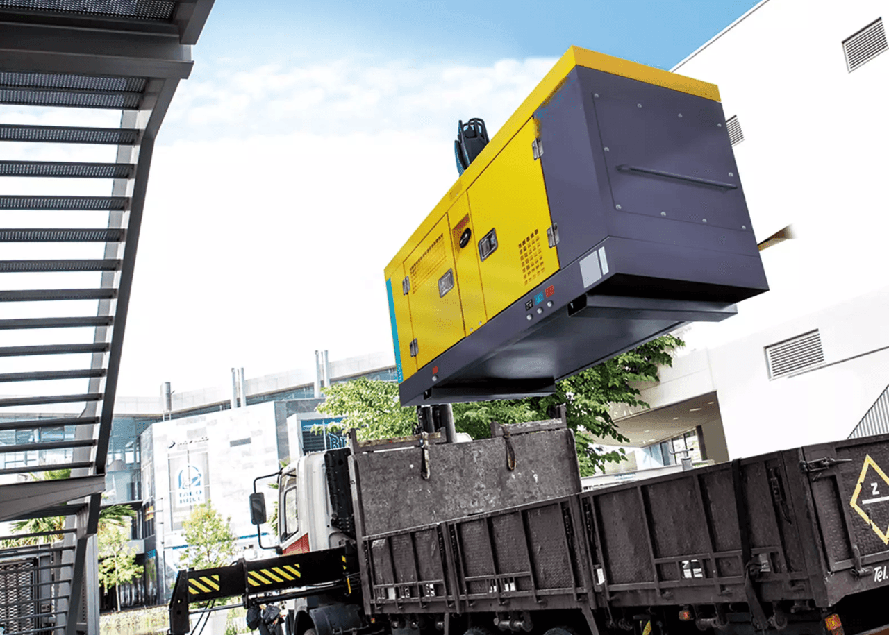 Generator being lifted by a crane onto a flatbed truck in an industrial area.